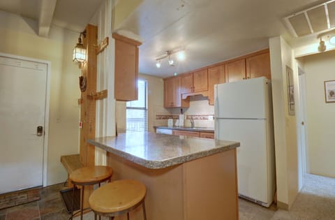 A kitchen featuring wooden cabinets, granite countertops, a white refrigerator, stove, and microwave. A small bar area with two wooden stools is in the foreground. The lighting is warm and inviting.