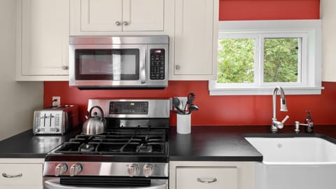 A modern kitchen features stainless steel appliances, including a stove, microwave, and toaster, with red backsplash, white cabinets, and a white sink under a window showing outdoor greenery.