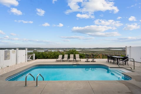 Private pool with lounge chairs, outdoor dining, white fencing, marsh and waterway views, blue sky, and relaxing coastal backdrop.