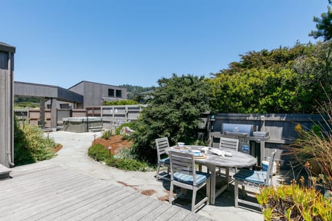 The private courtyard is designed for outdoor living, with a teak dining set, integrated grill, and adjacent hot tub arranged within a sheltered coastal setting.