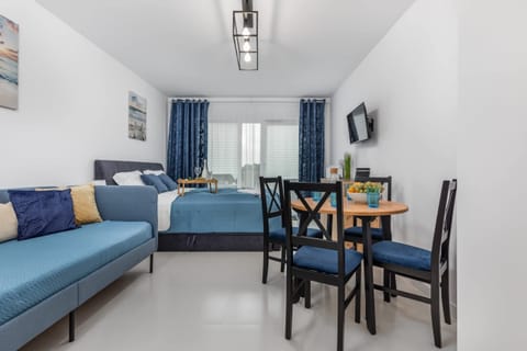A living room with a blue sofa and a dining area in the background, featuring a simple table and chairs, all styled in modern, neutral tones.