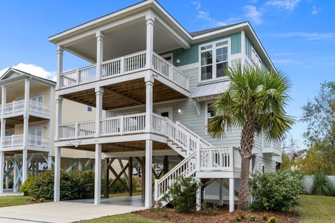 Elevated coastal home with two covered decks, white railings, exterior staircase, palm tree landscaping, and shaded parking beneath the house with concrete driveway in this Oak Island, NC vacation rental.