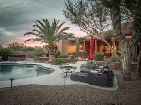 A serene desert retreat features a sparkling pool, hot tub, fire pit, and shaded loungers amid towering saguaros and lush palms. Sunset paints the sky as warm lights glow from the adobe-style home, creating a perfect Southwest oasis.