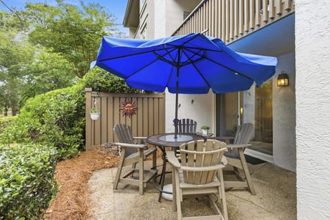 Vivid sapphire shades from the broad umbrella cast a refreshing glow over this stone patio. Weathered adirondack chairs gather around an intimate table, creating a peaceful corner for slow mornings and quiet conversation among the greenery.