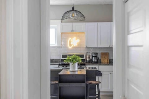 Charming kitchen with a central island featuring rustic stools and a leafy centerpiece. Behind, a neon “Café” sign glows above a sleek stove, flanked by white cabinets and a cozy coffee station. ️☕