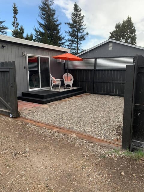 The seating area is accented by white wicker chairs, an orange patio umbrella, and a patterned rug, all set against a low-maintenance gravel yard.