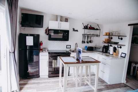 This modern studio kitchenette features clean white cabinetry, a marble-inspired countertop, and a versatile central island. Outfitted with a black refrigerator, gas range, and wall-mounted TV, it offers a stylish space.