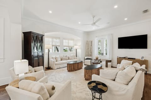 Neutral-toned seating surrounds two oval wood coffee tables beneath recessed lighting and a ceiling fan.