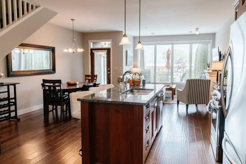 This bright open-concept kitchen and living area features a dark granite island, stainless steel appliances, and a cozy dining space. The striped armchair and large windows create a welcoming, airy atmosphere.