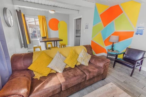 Vibrant and cozy living space featuring a warm brown couch with colorful pillows, a stylish geometric accent wall, and a bright dining area with yellow stools for a cheerful and inviting atmosphere.