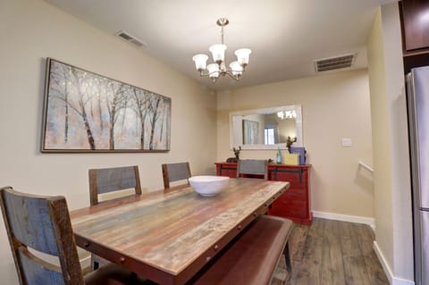 A dining room with a wooden table, six chairs, a red sideboard with decorative items, a framed tree artwork, and a rectangular mirror on the wall. A chandelier hangs above the table.