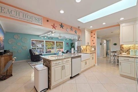 Open, airy coastal kitchen and living space filled with natural light. White cabinetry, granite countertops, skylights, and playful beach décor create a bright gathering spot perfect for cooking, relaxing, and enjoying time together by the water.