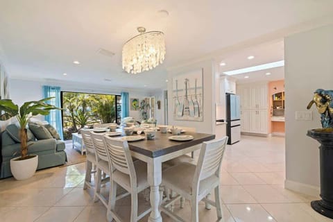 Open-concept dining and living space with coastal elegance. A large dining table invites gatherings beneath a shell-inspired chandelier, while bright décor, tropical views, and seamless flow to the kitchen create a welcoming, beachy atmosphere.