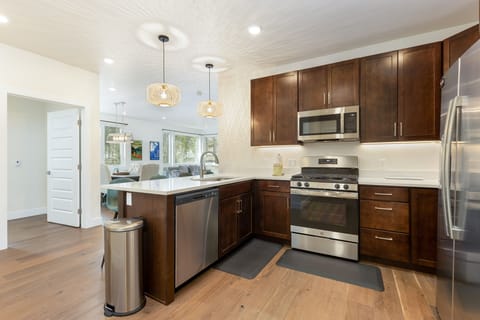 Modern kitchen with stainless steel appliances, dark wood cabinets, white countertops, and a wooden floor. A door to another room is visible in the background, along with dining area and hanging lights.