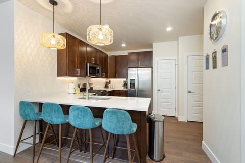 Modern kitchen with dark wood cabinets, stainless steel appliances, white countertops, and blue barstools. Pendant lights hang above the island with three stools. A trash bin is visible on the right.