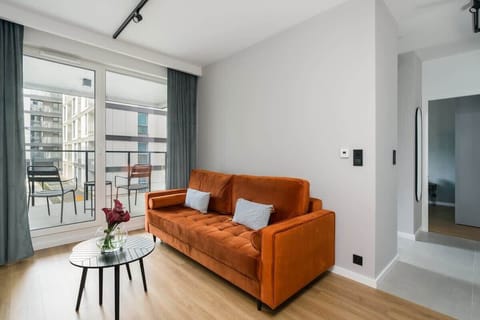 A bright and modern living room with a stylish orange couch, wooden floor, and large windows, allowing plenty of natural light.