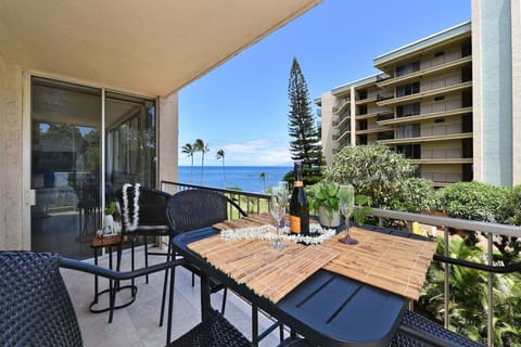 Balcony with table set for drinks, overlooking ocean views framed by palms and nearby building.