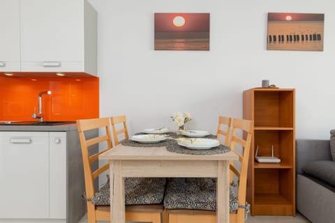 A dining area with a wooden table, chairs, and an orange kitchen backsplash. A small decorative shelf and framed artwork are also visible.