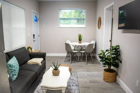 Living room with a comfortable sofa, coffee table, smart TV, and a dining area that seats four. Decorative plants add a fresh touch to the space.