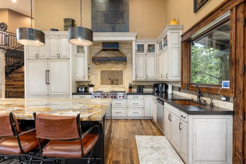 A spacious kitchen with white cabinets, a large island with a granite countertop, brown leather bar stools, stainless steel appliances, and a large window with a view of trees.