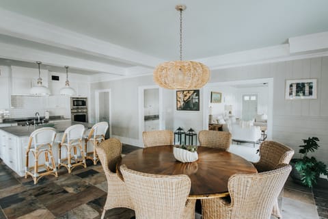 A spacious kitchen and dining area with wicker chairs, a round wooden table, hanging light fixture, and a view into a living room. White cabinets and bar stools are in the background.