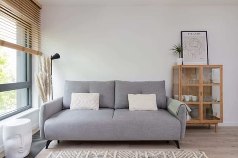 A cozy living room with a gray sofa, a wooden side table, and a minimalistic bookshelf, complemented by soft natural light from large windows.