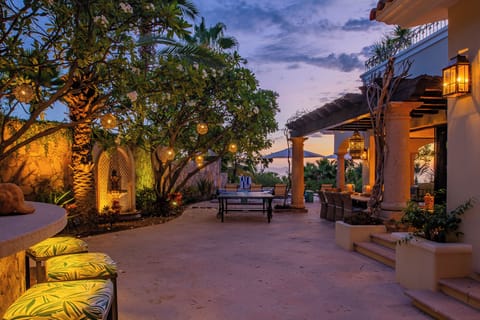 This outdoor courtyard glows as dusk settles, with lanterns casting warm light across inviting seating areas, lush palms, and serene architecture—an enchanting space for evening relaxation.