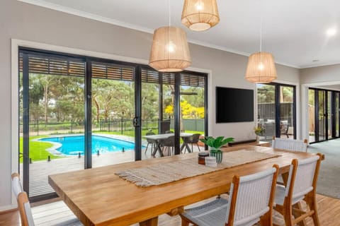 This bright and modern dining area features a wooden table, woven chairs, and pendant lights, with large glass doors opening to a backyard deck and a swimming pool, surrounded by trees for a peaceful, natural setting.