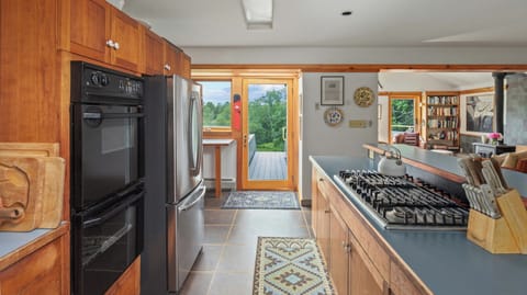 A kitchen with wooden cabinets, a black oven, and a stainless steel refrigerator. The countertop has a gas stove and a knife block. A rug is on the floor, and a doorway leads to a deck with greenery outside.