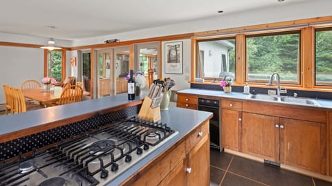 A kitchen with wooden cabinets, a gas stove, and a sink. Windows overlook a green yard. A dining area with a wooden table and chairs is visible in the background.