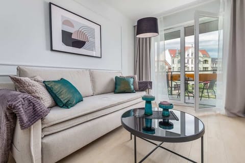 A stylish living room with a beige sofa, decorative pillows, a round coffee table, and a floor lamp, complemented by a balcony with a view of the city.