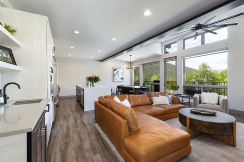 A modern living room with a brown leather sectional sofa, a round coffee table, and a kitchen in the background. Large windows offer a view of green trees and mountains. Ceiling fans and lights are on.