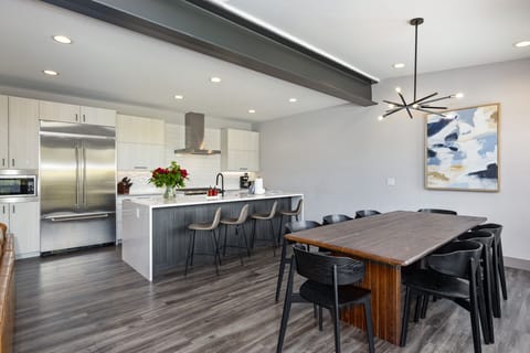 Modern kitchen and dining area with stainless steel appliances, a large island with bar stools, a wooden dining table with black chairs, and a contemporary pendant light.