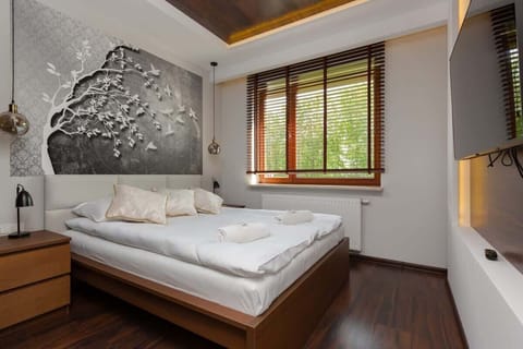 A modern bedroom featuring a double bed with white bedding, a wooden side table, and a large window letting in natural light.