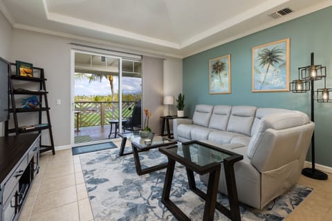 - Living room with plush sectional sofa glass coffee tables & tropical wall art accents - Sliding door opens to covered balcony with seating & views of palm lined landscape - Soft area rug and calming color palette create a relaxed welcoming atmosphere