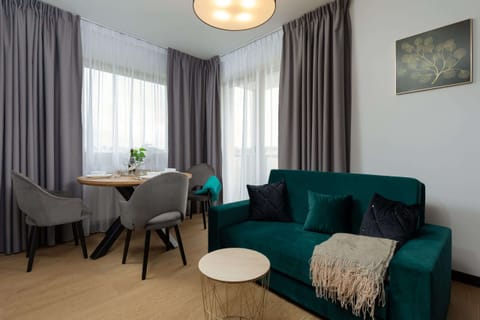 A wide shot showcasing the green sofa, dining table, and large window that lets in natural light, creating an airy atmosphere.