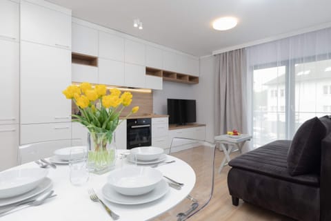 Close-up of a dining table with elegant white tableware, in the distance you can see a kitchenette, a TV and sofa.