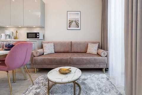 A bright living room with a pink sofa, round coffee table, and white furniture, complemented by a large window.
