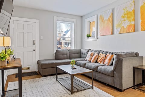 Front living room view with a gray L-shaped sectional, orange accent pillows, coffee table, wall-mounted TV and white entry door.