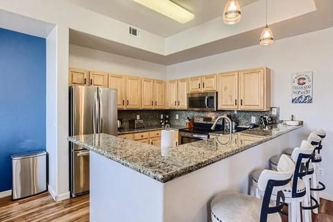 Modern kitchen featuring wooden cabinets, granite countertops, stainless steel appliances, and a breakfast bar with three stools. Pendant lights hang above the bar. A blue accent wall is visible.