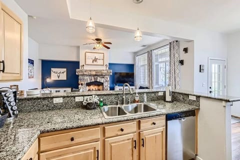 A bright kitchen with light wooden cabinets, granite countertops, and double sink. The kitchen opens to a living area featuring a fireplace and a ceiling fan.