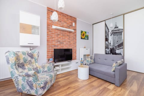 A cozy living space featuring a dark gray sofa, a glass coffee table, and a decorative brick wall. The room is complemented by a large plant and framed art.