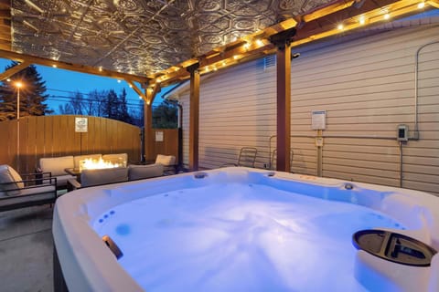 Steaming hot tub under a covered patio strung with warm lights, creating a cozy spot to sink in and unwind. Lounge seating and wood fencing frame the space, inviting slow evening soaks and quiet conversations outdoors.