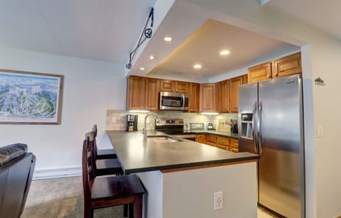 A modern kitchen features wooden cabinets, stainless steel appliances, a double sink, and a countertop with bar stools. A wall painting and recessed lighting enhance the space.
