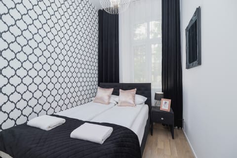 A bright bedroom with a double bed, patterned wallpaper, and a large window with dark curtains. Decorative pillows add a cozy touch to the interior.