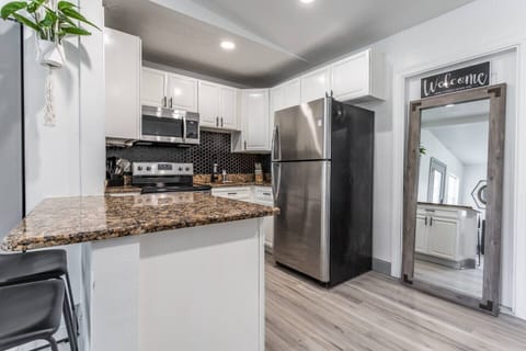 A modern, fully equipped kitchen featuring sleek white cabinetry, stainless-steel appliances, and a stylish black backsplash. The granite countertops add a touch of elegance and provide ample workspace for meal preparation. A "Welcome" sign above a full-length mirror adds warmth and charm to the space. A hanging plant introduces a hint of greenery, while light wood flooring cre...