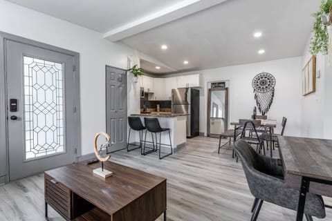 Living room and kitchen: An open-concept living space with a modern and inviting design. The room features a sleek wooden coffee table in the foreground and a stylish work desk and chair on the right, ideal for a workspace. The kitchen area, with its white cabinetry and stainless-steel appliances, includes a breakfast bar with high stools for casual dining. A glass-paneled fron...