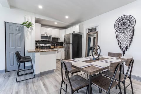 A modern, cozy dining and kitchen area featuring a wooden dining table with seating for six, accented by a decorative centerpiece. On the wall, a stylish dreamcatcher adds a unique touch. The kitchen is equipped with white cabinetry, stainless-steel appliances, and a sleek black backsplash. A breakfast bar with two high stools offers additional seating. Light wood flooring and ...