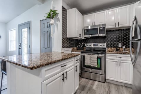 A modern, fully equipped kitchen featuring sleek white cabinetry, stainless-steel appliances, and a stylish black backsplash. The granite countertops add a touch of elegance and provide ample workspace for meal preparation. A "Welcome" sign above a full-length mirror adds warmth and charm to the space. A hanging plant introduces a hint of greenery, while light wood flooring cre...