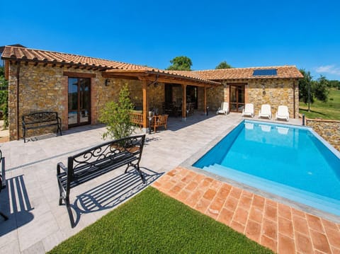 A rustic stone villa with a terracotta roof features a covered patio and a sparkling blue swimming pool. White lounge chairs and a black metal bench sit on a light grey tiled patio under a clear blue sky.